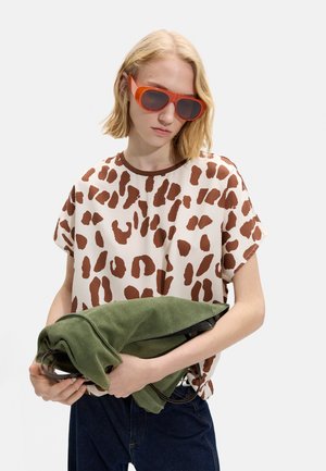 Kobieta w pomarańczowych okularach przeciwsłonecznych i białej koszuli w brązowe plamy, trzymająca złożoną zieloną kurtkę na jednolitym białym tle.