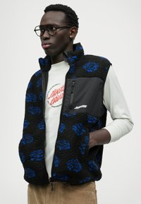 Fekete és kék mintázatú bolyhos mellény, cipzáras fekete anyagú mellszárral, világos színű ing fölött viselve. Kerek formákkal díszítve.