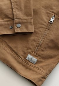 Zbliżenie na brązowy rękaw kurtki z tkaniny z metalowym zatrzaskiem, ukośną zamkniętą zamkiem kieszenią oraz białą prostokątną etykietą marki z tkaniny.