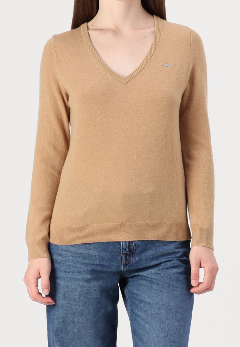 Femme portant un pull col V couleur chameau et un jean bleu, debout les mains détendues le long du corps sur un fond uni.