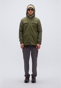 Πράσινο ελαιολαδί μπουφάν με κουκούλα και δύο τσέπες στο στήθος, συνδυασμένο με γκρι παντελόνια και σκούρα παπούτσια. Το μοντέλο φορά γυαλιά ηλίου και ελαφρύ σκουφάκι.