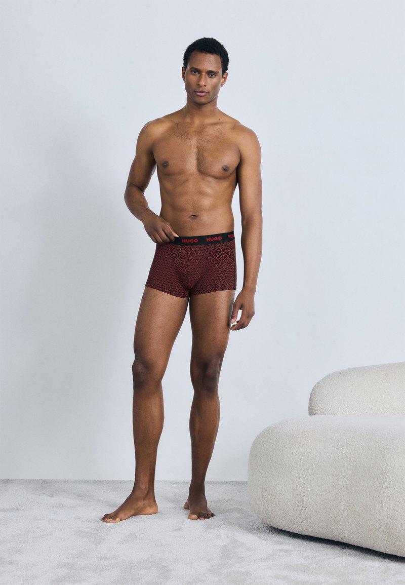 Cuecas boxer vermelhas com padrão, cós preto com o logótipo "HUGO" em vermelho. O tecido parece macio e ajustado ao corpo.