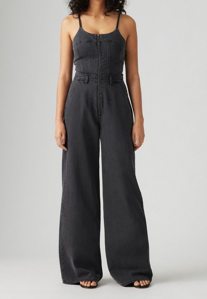 Femme portant une combinaison en denim foncé sans manches, à fines bretelles, fermeture zippée devant, jambes larges et sandales ouvertes, debout.