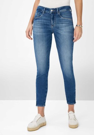 Femme portant un jean skinny bleu taille mi-haute et des chaussures blanches à lacets, debout avec les bras détendus le long du corps, sur un fond clair uni.