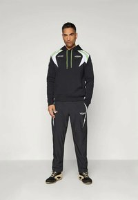 Crni trenirku s bijelim i zelenim detaljima. Dukserica ima patentni zatvarač i logo, a hlače su sužene prema nogavicama. Tkanina izgleda glatko i sportski.