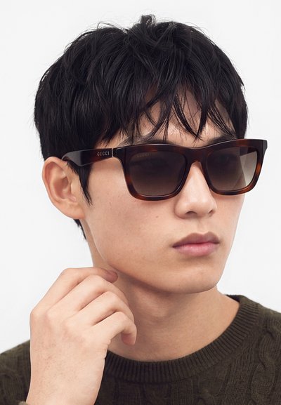 Ung mand med kort sort hår iført mørke, rektangulære Gucci-solbriller og en grøn strikket sweater, der holder sin hånd tæt på sin hage.