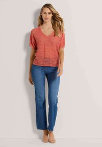 Rotes-orange gestricktes Oberteil mit V-Ausschnitt und lockerer Passform, kombiniert mit blauen Jeans im Straight-Leg-Schnitt und transparenten Absatzsandalen. Glatte Textur, lässiges Design.