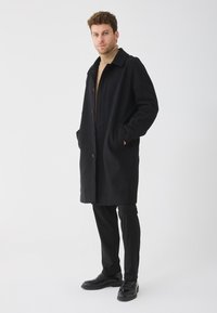 Abrigo de lana negra con cuello, botones frontales y bolsillos laterales, combinado con pantalones negros ajustados y zapatos pulidos.