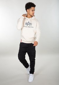 Felpa beige con logo grigio scuro, dotata di tasca frontale; abbinata a pantaloni cargo neri e sneakers bianche. Outfit semplice e casual.