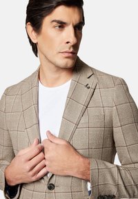 Beżowa marynarka w kratę z teksturowanym wzorem, brązowymi akcentami, zapięciem na dwa guziki oraz jedną kieszenią. Nosi się ją na białej koszulce.