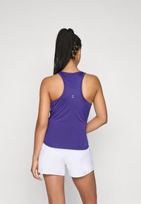 Lila sporttopp med racerbackdesign, slät textur och en liten vit logodetalj på baksidan; kombinerad med vita shorts.