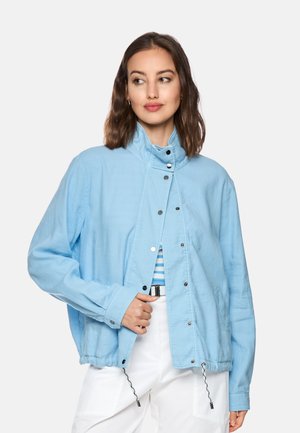 Woman wearing light blue buttoned jacket over striped top and white pants, looking to her left with a slight smile.