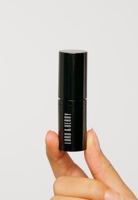 Zwarte cilindrische lippenstift tube met zilveren accenten, gemarkeerd met "LORD & BERRY" in witte tekst, vastgehouden tussen vingers tegen een effen achtergrond.