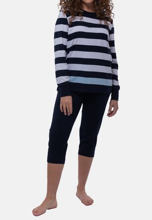 Vrouw draagt een longsleeve met zwart-witte horizontale strepen en zwarte cropped broek, staat blootsvoets tegen een effen achtergrond.