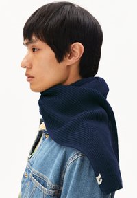 Écharpe texturée navy drapée sur l'épaule d'un mannequin, avec un design à côtes et une petite étiquette blanche sur le bord. Veste en denim en dessous.