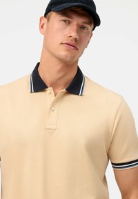 Beige polo shirt met een gestructureerde afwerking, voorzien van een marineblauwe kraag en gestreepte accenten. Twee-knopen knoopsluiting met een klein logo op de borst.