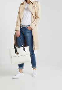 Bílá taška přes rameno s černými madly a stužkovými akcenty, s ručně psaným logem. Modelka má na sobě béžový trenčkot, bílý tričko a džíny.