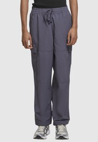 Personne portant un pantalon cargo ample gris foncé avec une taille à cordon et des chaussures de course blanc-gris sur un fond clair uni.