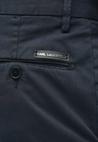 Calças azul-marinho feitas de tecido suave. Apresentam uma placa com logótipo preto, fecho com botão e um pequeno bolso lateral.