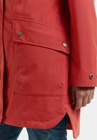 Rød vandtæt jakke med store klaplommer og metaltrykknapper, båret over blå denimjeans med synligt sidelomme og ærmekant.