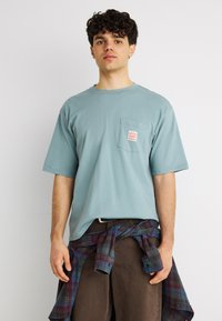 Νεαρός με σγουρά μαλλιά που φοράει ένα ανοιχτό μπλε φαρδύ μπλουζάκι, με καρό πουκάμισο δεμένο στη μέση και καφέ παντελόνι, μπροστά σε απλό φόντο.