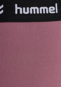 Nærbillede af sort taljebånd med hvid "hummel" logo over tekstureret malvafarvet stofpanel.