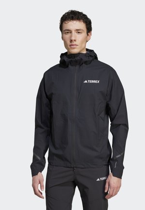 Hombre con chaqueta impermeable y pantalones negros Adidas Terrex, de pie frente a un fondo claro liso, mirando al frente con los brazos relajados.