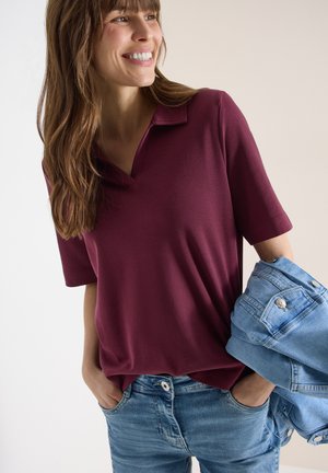 Lächelnde Frau mit einem kurzärmligen Poloshirt in Burgunderrot und hellblauer Jeans, die eine gefaltete Jeansjacke in der linken Hand hält.