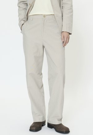 Egy férfi bézs tűsarkú nadrágot, bézs blézert, fehér pulóvert és sötétbarna bőr cipőt visel, egy egyszerű fehér háttér előtt.