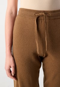 Pantaloni in maglia marroni con una texture morbida, vita alta e coulisse regolabile. Il primo piano mostra la vita e una mano che la tiene.