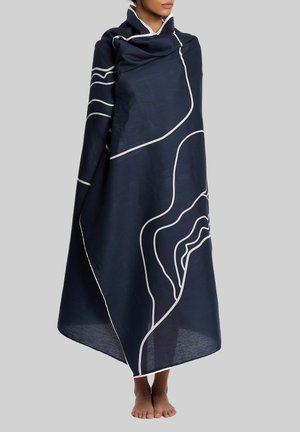 Persona envuelta en un gran chal azul marino con líneas de contorno abstractas blancas, de pie descalza sobre un fondo sencillo.