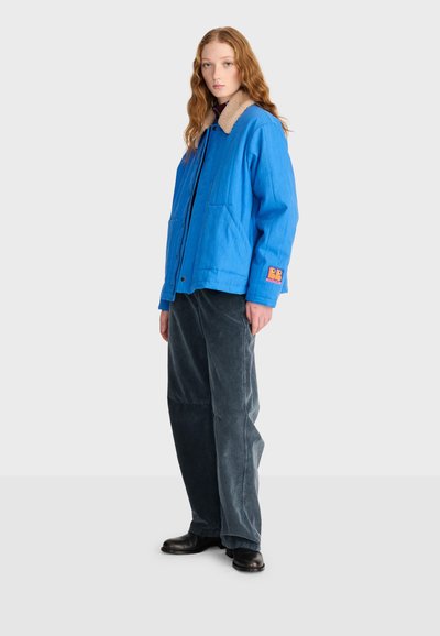 Jeune personne aux longs cheveux roux debout, portant une veste bleue à col beige, un pantalon ample foncé et des chaussures noires sur un fond uni.