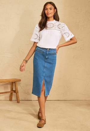 Kvinde stående indendørs iført en hvid øjletsbluse og en blå denim midi nederdel med frontslids og brune sko.