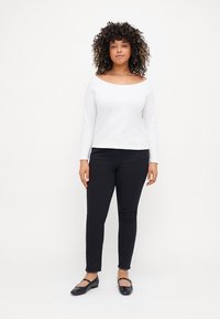 Camiseta blanca de manga larga con escote露hombros, combinada con pantalones vaqueros ajustados negros y zapatos planos negros, sobre un fondo liso.