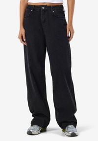 Crne traperice širokih nogavica, izrađene od trapera, s visokim strukom, prednjim džepovima i blago izblijedio završenim izgledom. Uparene sa srebrnim tenisicama.