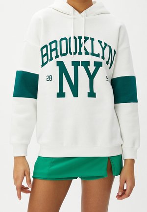 Bílá mikina s zeleným nápisem „BROOKLYN NY“ a zelenými pásky na rukávech, nošená se zelenou mini sukní s předním rozparkem.
