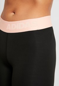 Černé sportovní legíny se širokým růžovým pasem s nápisem „ADIDAS“ v texturovaném tónovaném potisku. Hladká látka, přiléhavý střih.