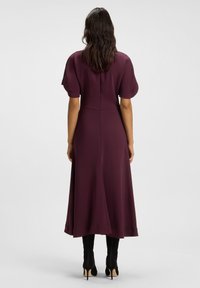 Donna con lunghi capelli mossi indossa un vestito midi viola con maniche corte e stivali neri con tacco, in piedi di spalle su uno sfondo neutro.