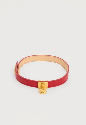 Rødt læder choker-halskæde med gyldent firkantet spænde og rund gylden vedhæng med bogstavet "C" på hvid baggrund.