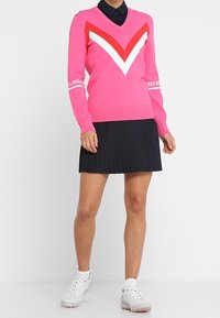 Maglione rosa a V con motivo a zigzag bianco e rosso; abbinato a una gonna plissettata nera e scarpe da ginnastica bianche, con colletto.