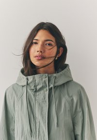 Light green, hooded jacket made of crinkled fabric. Features drawstrings, minimalist snap buttons, and a relaxed fit design.