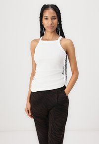White ribbed tank top with wide straps, paired with brown pants featuring a black tiger stripe pattern. Model is standing with hands in pockets.