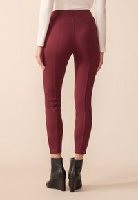 Pantalons ajustés bordeaux avec taille haute, présentant des détails de coutures et une texture lisse, associés à des bottines à talons compensés noires.