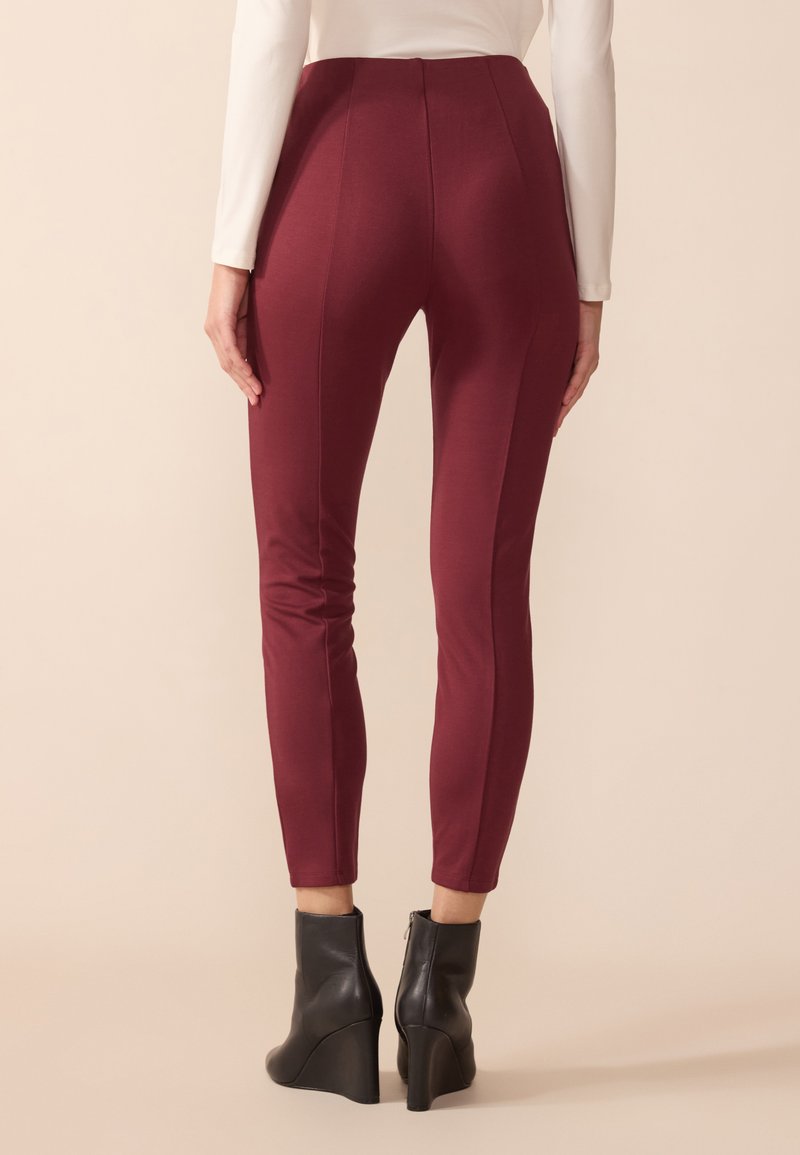 Pantaloni aderenti bordeaux a vita alta, con dettagli delle cuciture e una texture liscia, abbinati a stivaletti con tacco a zeppa neri.