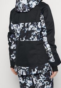 Bakifrån på en person som bär en svart jacka med blå och vita blommönster på huvan, baksidan, ärmarna och matchande byxor.