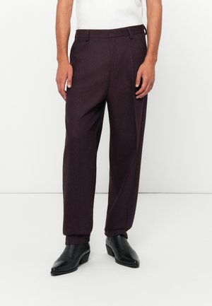 Man wearing dark purple tailored trousers and black leather ankle boots, standing against a plain white background.