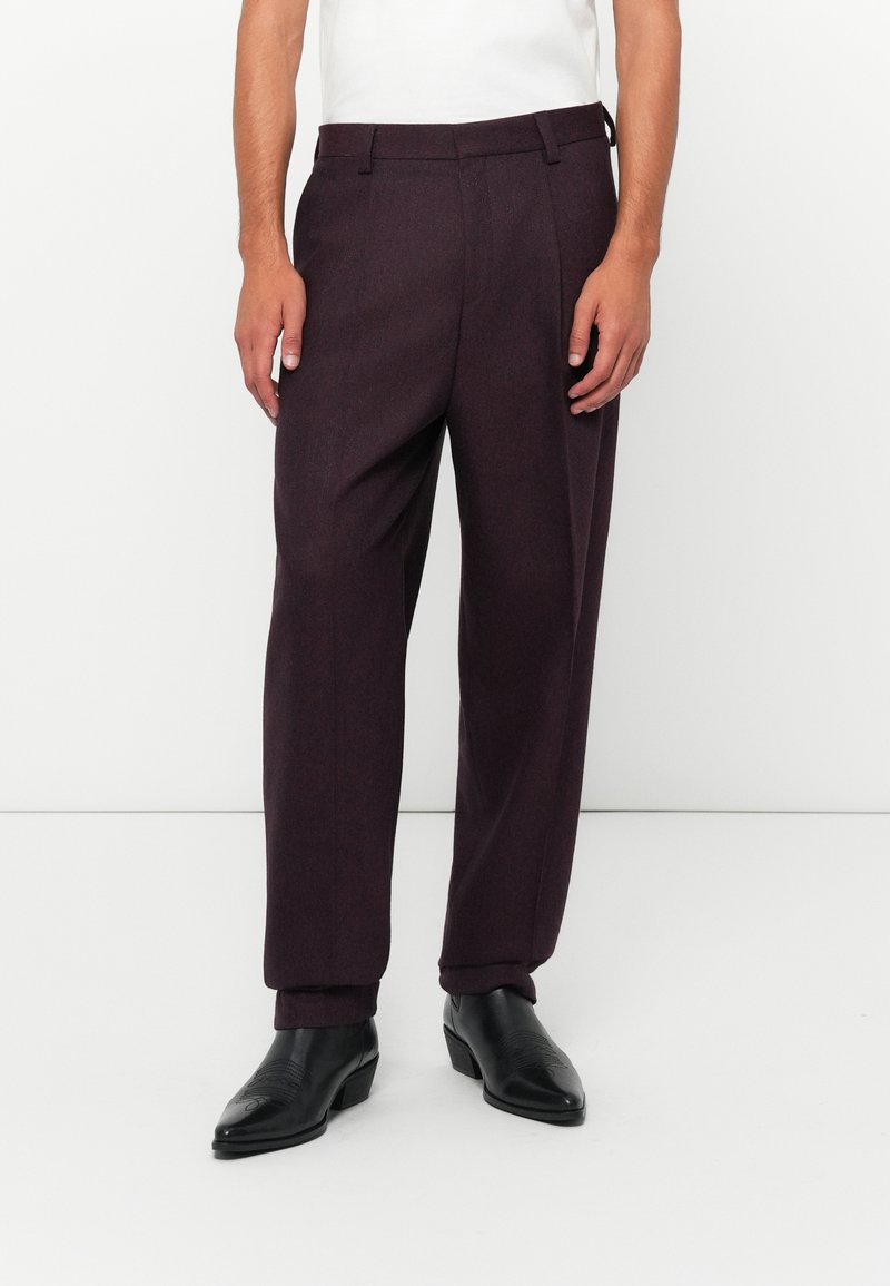 Man wearing dark purple tailored trousers and black leather ankle boots, standing against a plain white background.