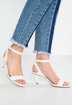 Weiße High-Heel-Sandaletten mit minimalistischem Design, bestehend aus zwei breiten Riemen und einem Knöchelriemen, kombiniert mit verwaschenen blauen, cropped Jeans.