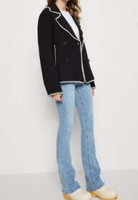 Zwarte double-breasted jas met contrasterende witte stiksels, gecombineerd met lichtblauwe flared jeans en crèmekleurige clogs.