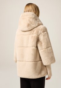 Beżowa kurtka ze sztucznego futra z dużym kapturem, krótkimi rękawami i poziomymi szwami, o miękkiej fakturze i swobodnym kroju.